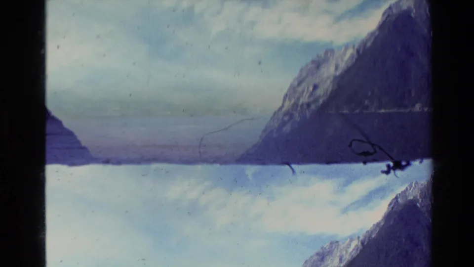 1981 New Zealand home-movie frame with a steep mountain face, water at the base, and visible 8mm scratches across the scene.