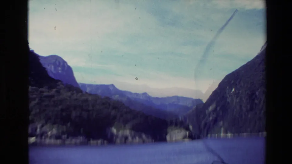 Wide 1981 New Zealand 8mm frame showing blue water, dark foreground slopes, and layered mountain ridges under a cloudy sky.