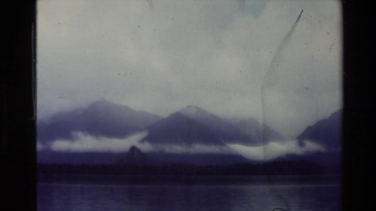 1981 New Zealand 8mm view of a calm lake, dark shoreline, distant mountains, and low cloud bands under a pale overcast sky.