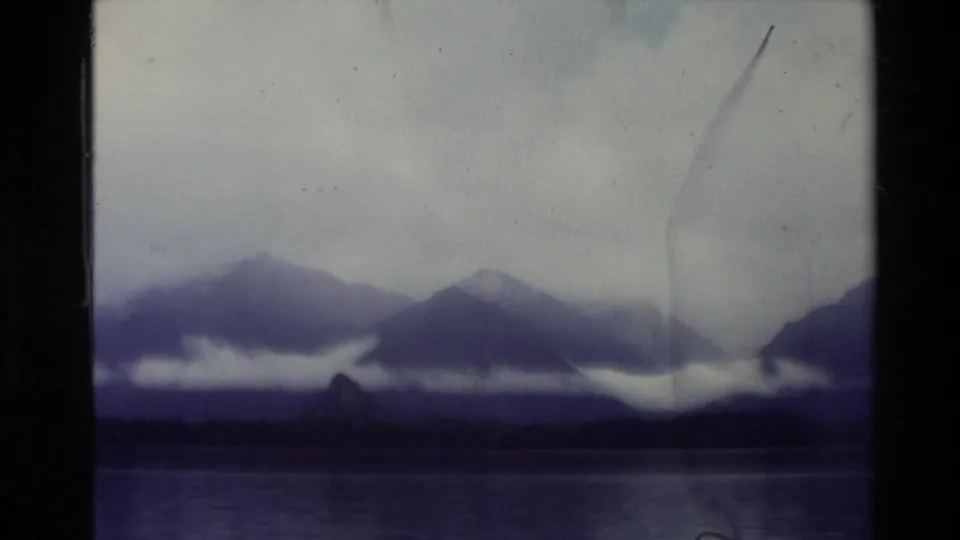 1981 New Zealand 8mm view of a calm lake, dark shoreline, distant mountains, and low cloud bands under a pale overcast sky.
