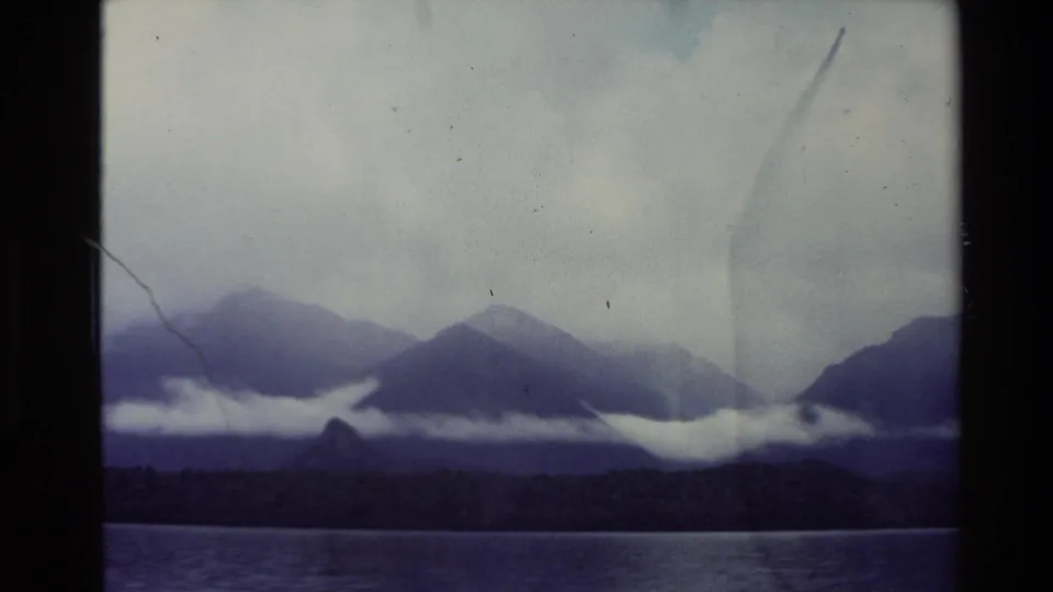 1981 New Zealand 8mm landscape showing a lake foreground, dark tree-lined shore, layered mountains, and low white clouds across the slopes.