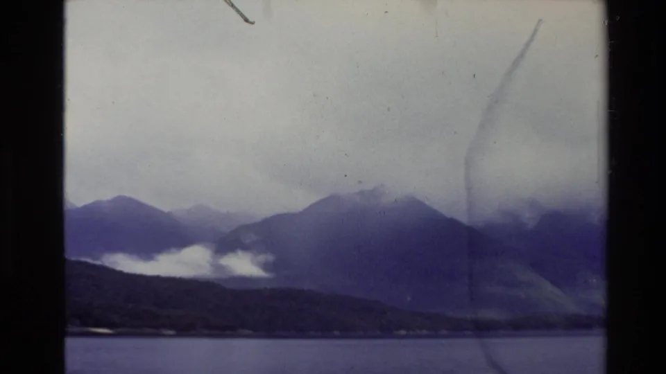 1981 New Zealand 8mm frame with still water, a dark shoreline, blue-purple mountain silhouettes, low clouds, and visible film wear.