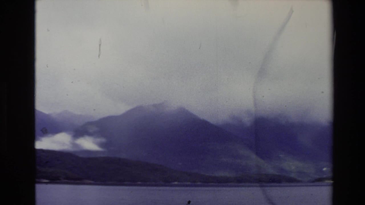 1981 New Zealand 8mm frame showing cloud-covered mountains beyond calm water, tinted purple with film grain and scratches.