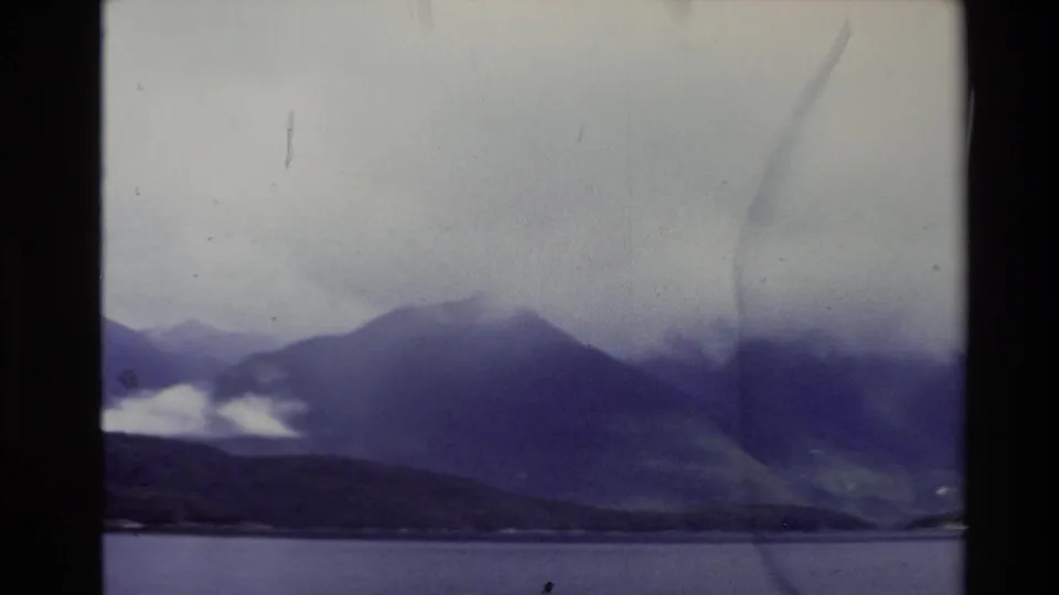 1981 New Zealand 8mm frame showing cloud-covered mountains beyond calm water, tinted purple with film grain and scratches.