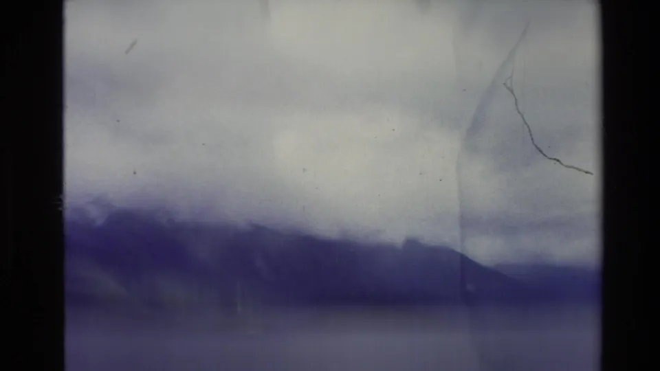 Soft 8mm view of New Zealand mountains across water, with cloud cover, purple cast, grain, and visible surface scratches.