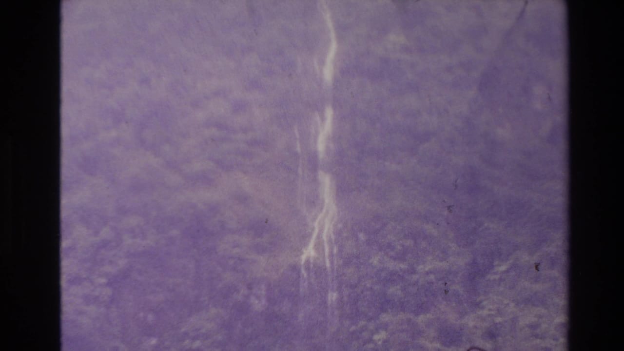 1981 New Zealand 8mm frame showing faint cascading water through purple color shift, grain, scratches, and dark frame borders.