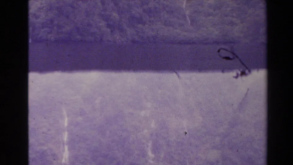 New Zealand 1981 8mm frame showing a distant waterfall area above pale water or mist, with purple fading, scratches, and dust marks.