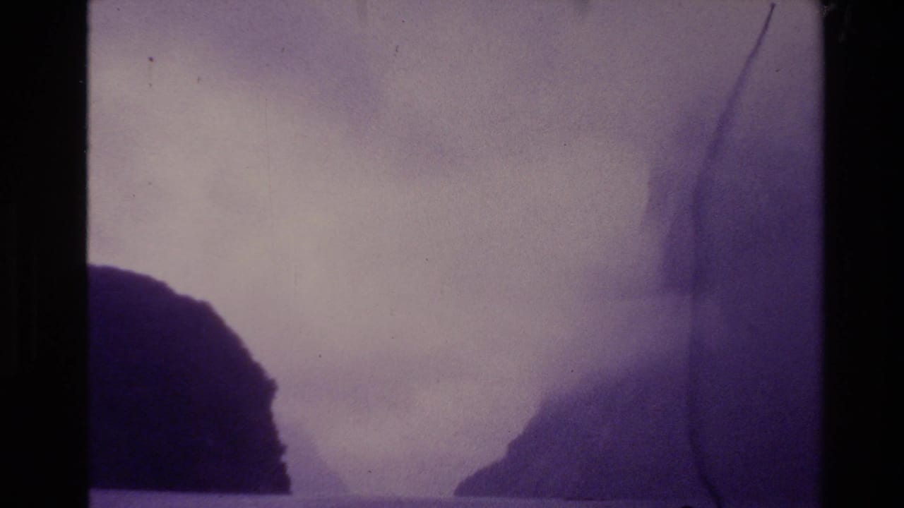 1981 New Zealand home-movie frame with fog separating dark hills across water, seen through grain and edge wear.