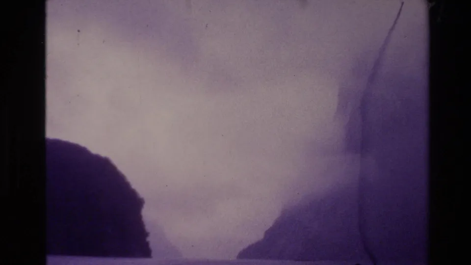 1981 New Zealand home-movie frame with fog separating dark hills across water, seen through grain and edge wear.