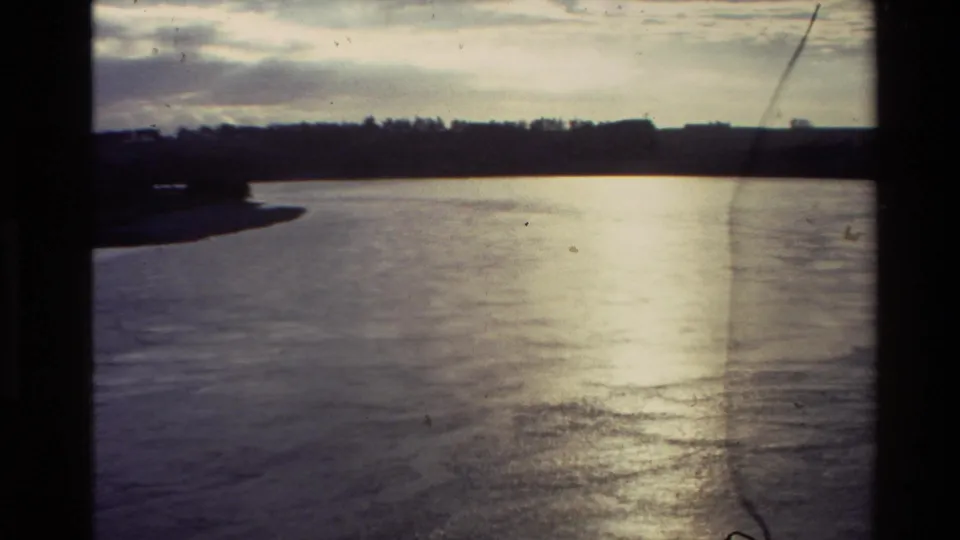1981 New Zealand 8mm view of a broad river bend, forested far bank, cloudy sky, and sunlit current.