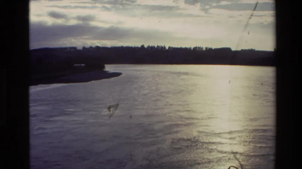 New Zealand 1981 home-movie frame showing a river bend, distant tree line, cloudy sky, and strong glare on moving water.