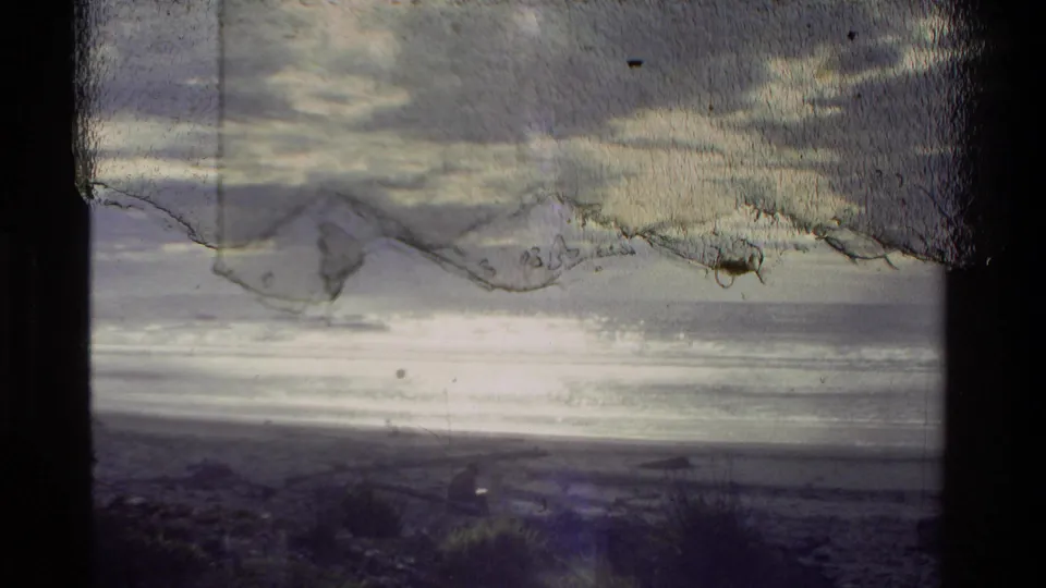 1981 New Zealand 8mm frame with a low river or shore horizon, cloud layers, bright water reflection, and pronounced surface wear.