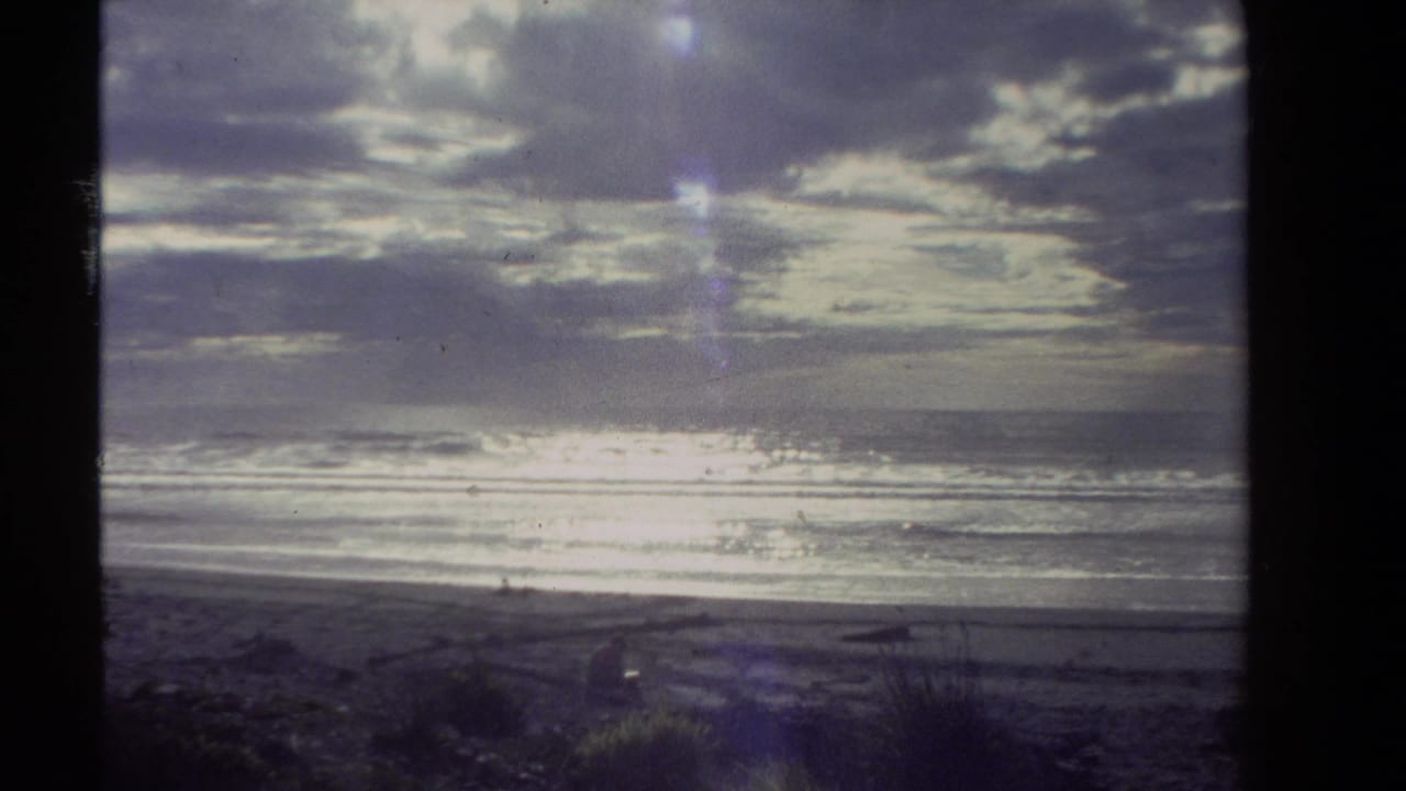 1981 New Zealand 8mm frame showing a broad beach, low surf, heavy clouds, and bright glare across the sea.