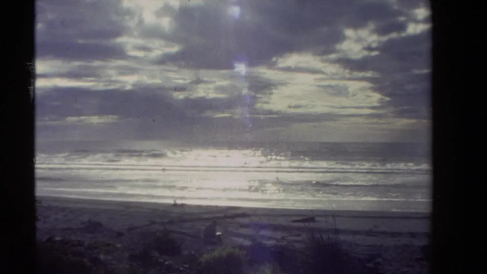 1981 New Zealand 8mm frame showing a broad beach, low surf, heavy clouds, and bright glare across the sea.