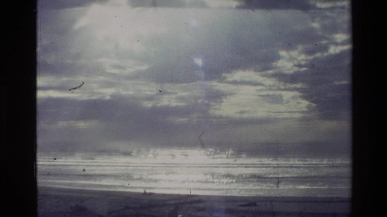1981 New Zealand 8mm frame of a coastal horizon with textured clouds, shimmering water, and a dark shoreline.