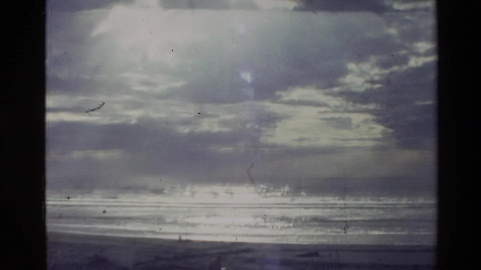 1981 New Zealand 8mm frame of a coastal horizon with textured clouds, shimmering water, and a dark shoreline.