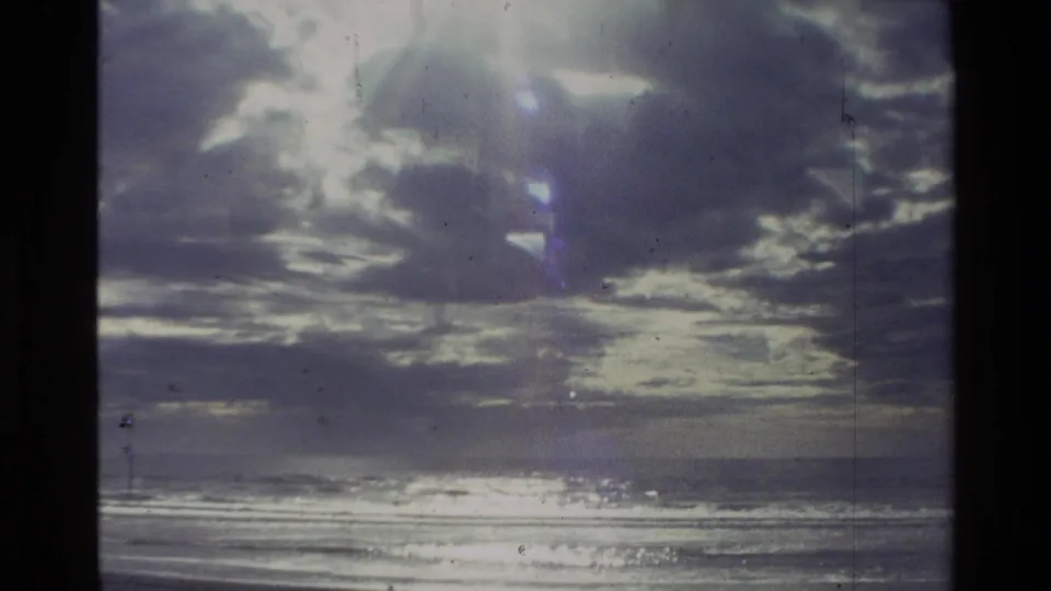 1981 New Zealand 8mm frame showing layered clouds, a reflective ocean edge, and an empty beach seen from a home-movie viewpoint.