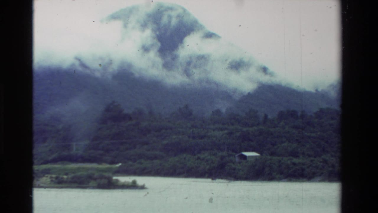 1981 New Zealand 8mm frame showing a cloud-wrapped forested mountain, lake water, and a small building near the shoreline.