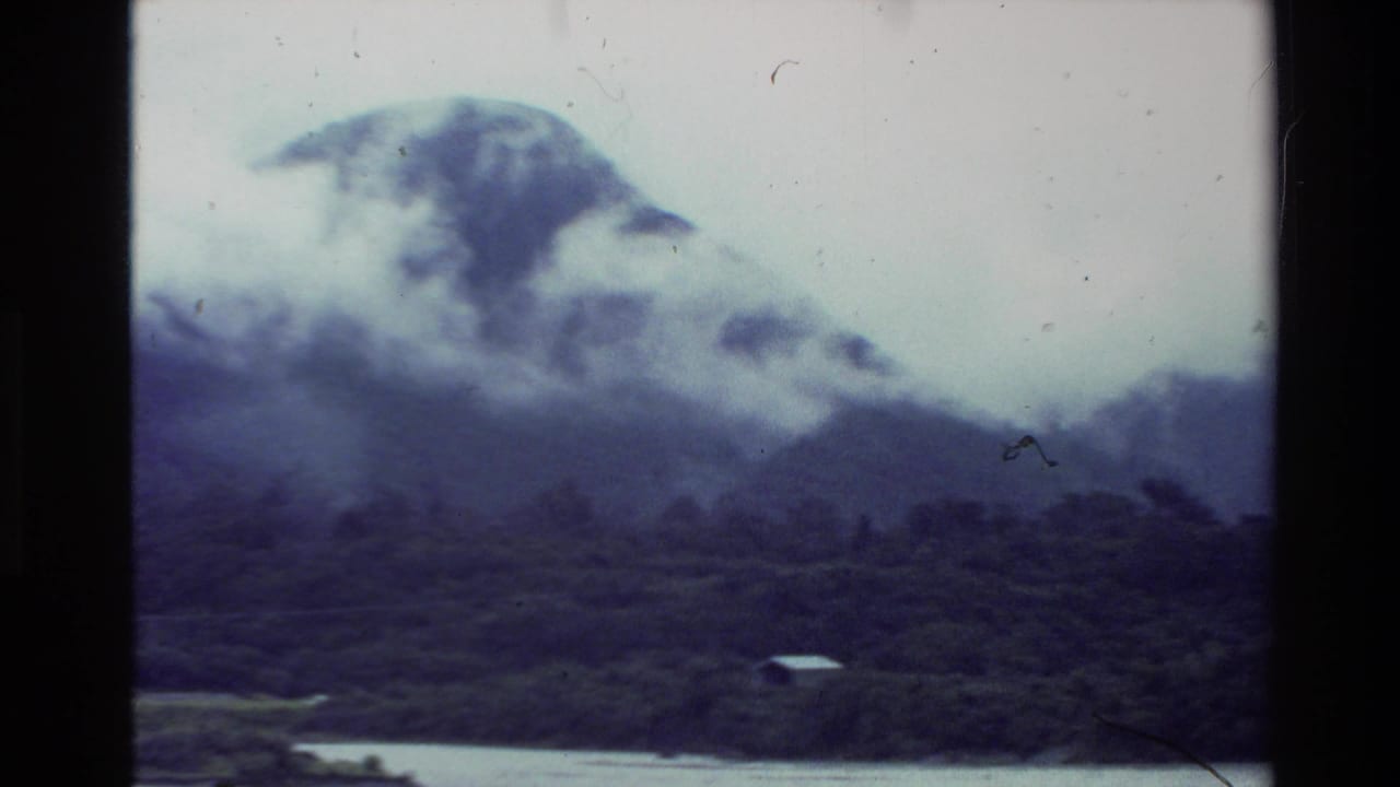 1981 New Zealand 8mm view from across water, with fog moving around a forested mountain and a small shoreline structure below.