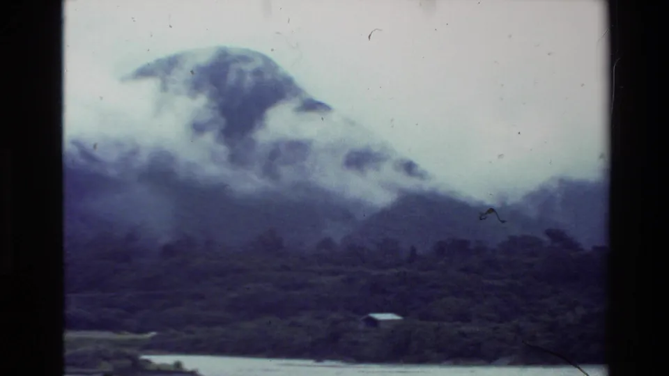1981 New Zealand 8mm view from across water, with fog moving around a forested mountain and a small shoreline structure below.