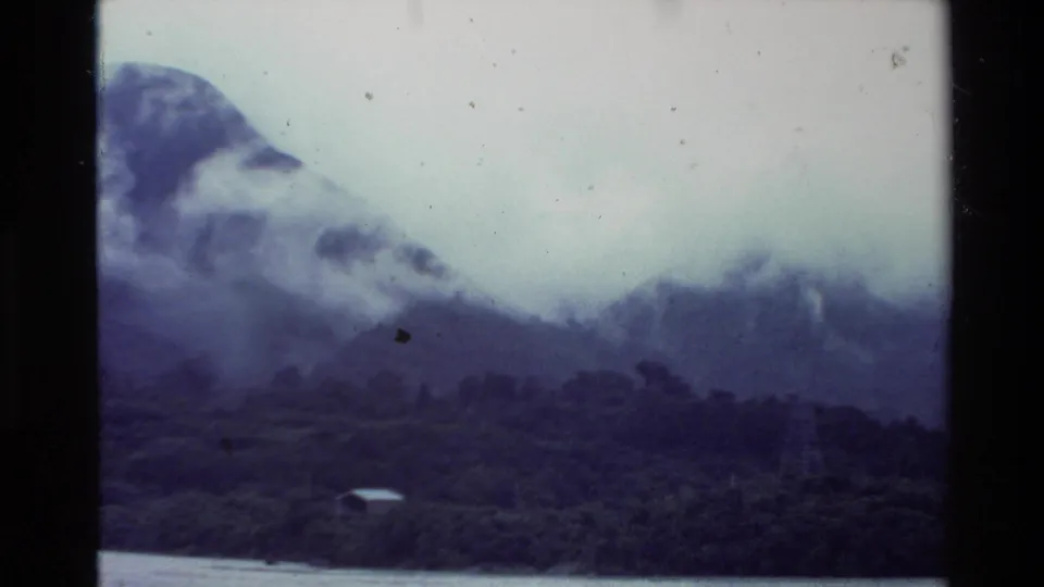 1981 New Zealand home-movie frame with a misty mountain slope, dense trees, lake water, and a small shoreline building.