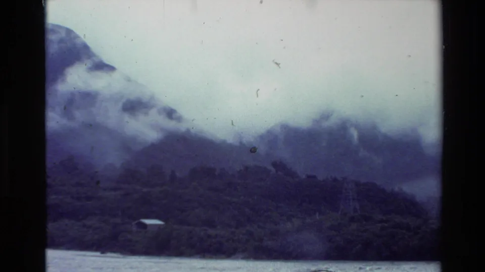 1981 New Zealand 8mm landscape frame showing a fog-softened mountain ridge, wooded shoreline, lake water, and visible film borders.