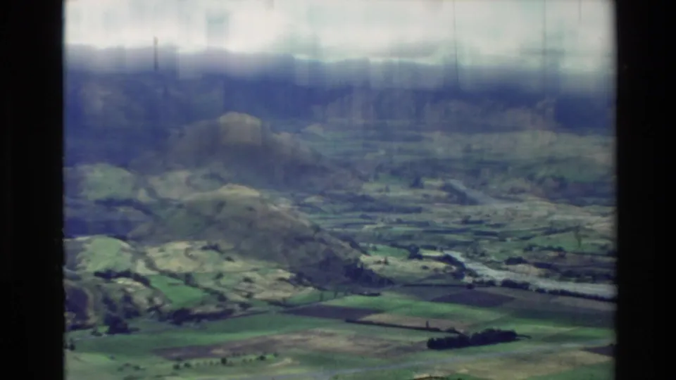 1981 New Zealand 8mm frame with rolling green countryside, valley fields and a prominent rounded hill below a cloudy sky.