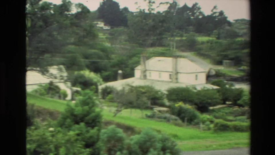 1981 New Zealand 8mm frame of a white rural house set among green grass, shrubs, and trees on a hillside.