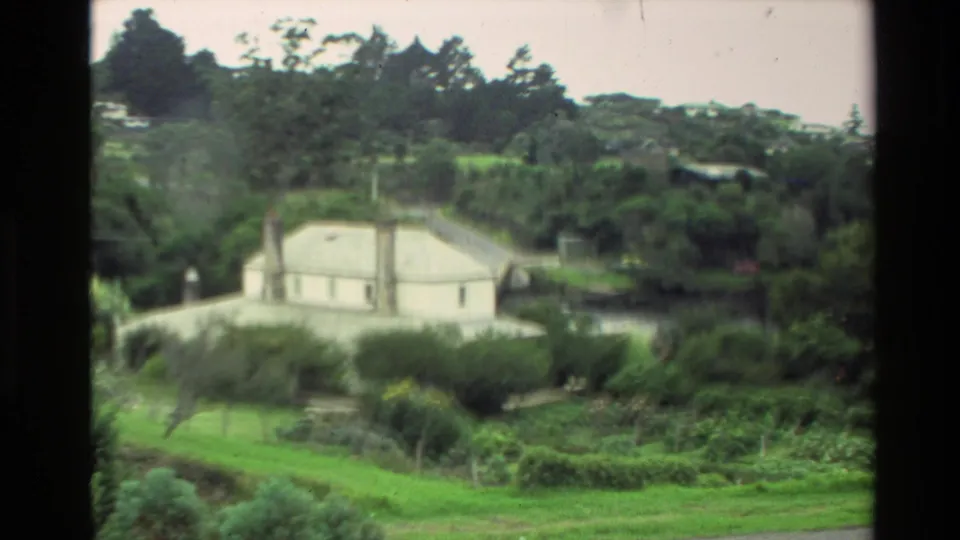 Soft 1981 New Zealand 8mm view of a white house in a bushy green area, with trees and grass filling the frame.