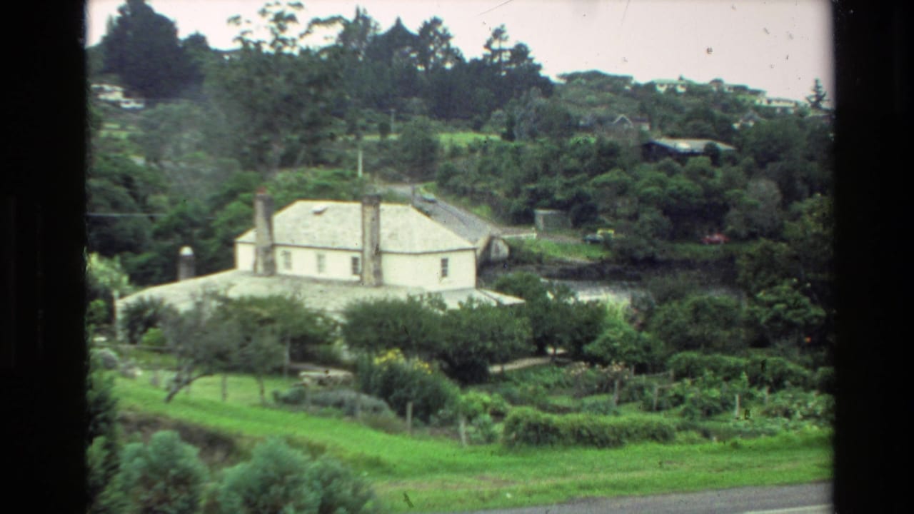 1981 New Zealand home-movie frame of a white house in a wooded, hilly setting with garden rows and a dark frame edge.