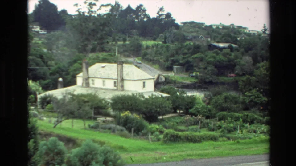 1981 New Zealand 8mm frame with a white house, wooded slope, garden foreground, and visible film-frame edges.