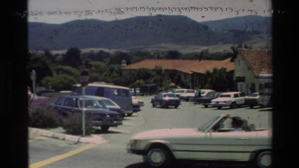 Carmel-area parking lot in 1982 with parked cars, a van, low red-tile buildings, trees, and rolling hills beyond.