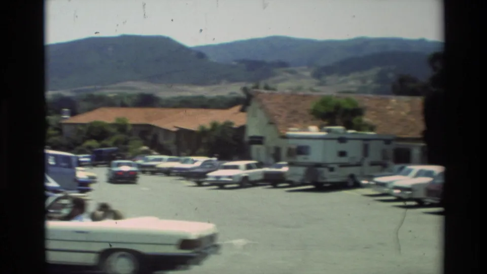 1982 Carmel-area parking lot view with a white camper trailer, parked cars, tile-roof buildings, trees, and hills behind.