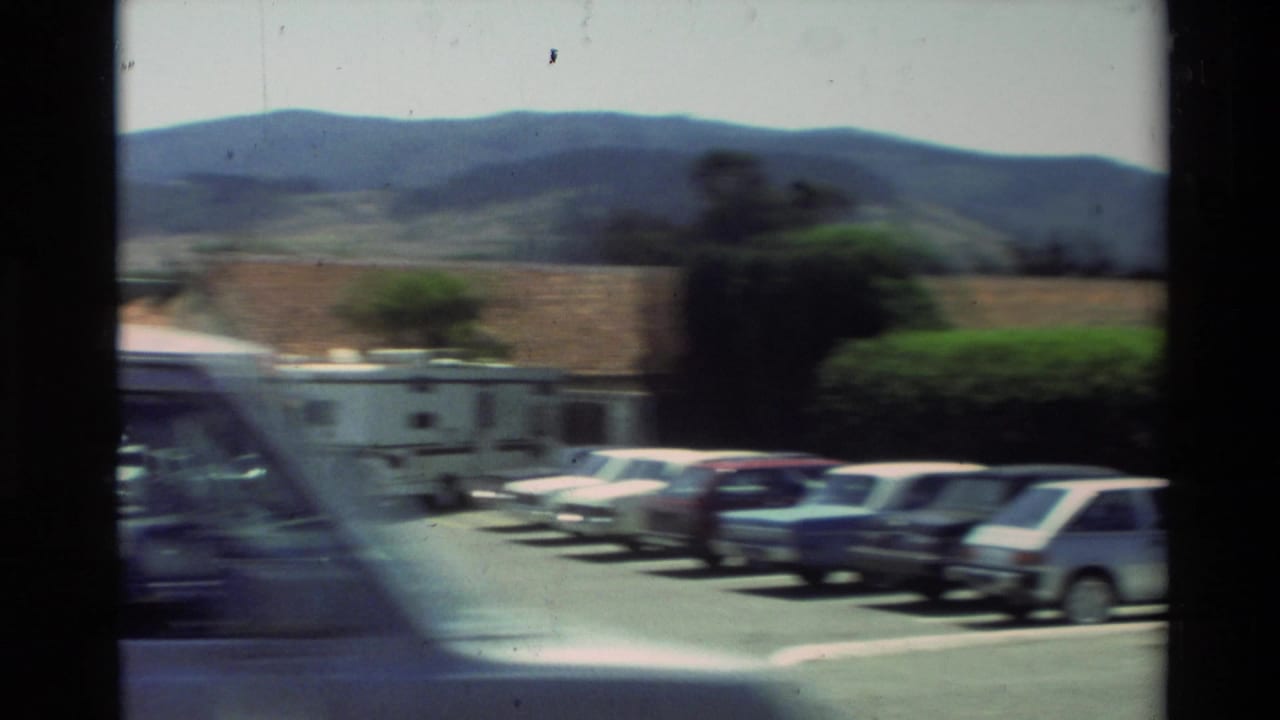 Carmel, California home-movie view of parked cars near Carmel Mission Basilica with hills behind the grounds.