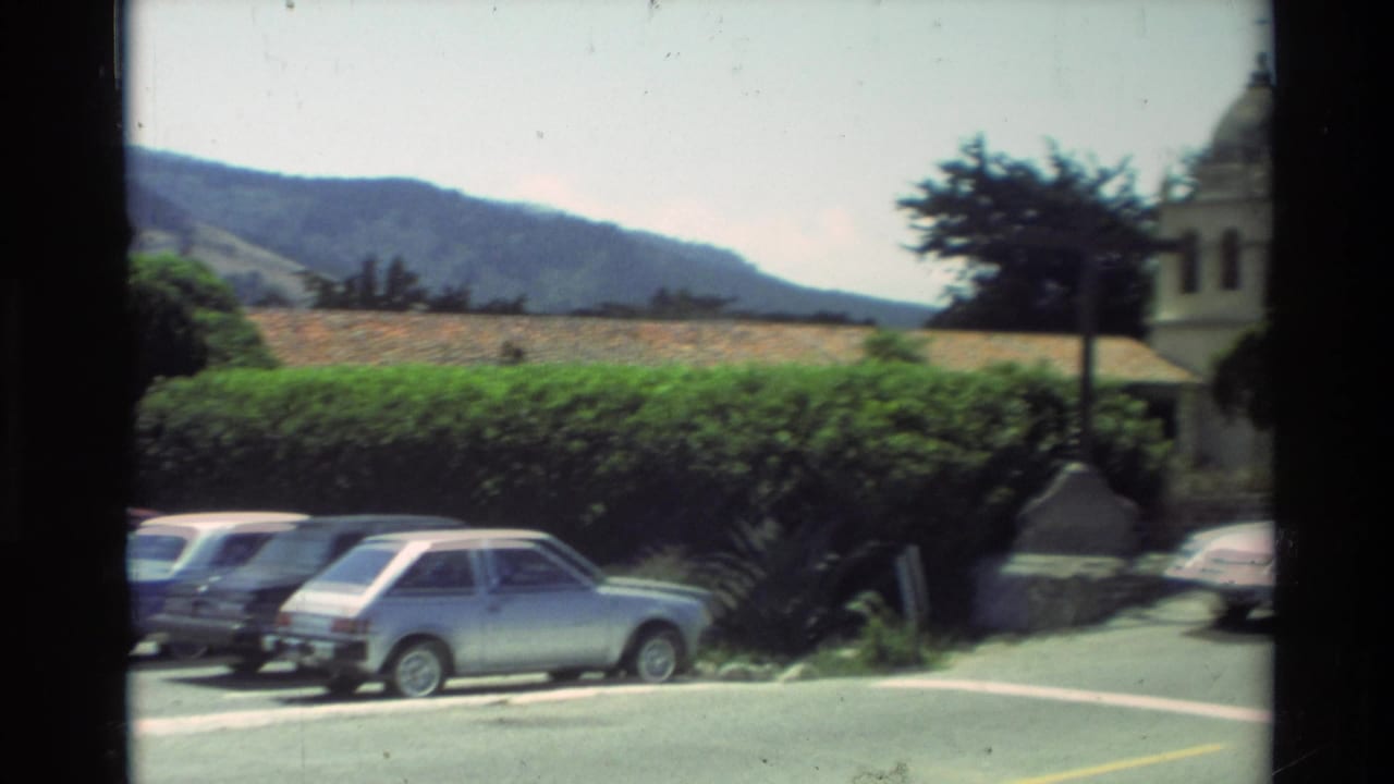 Cars parked beside hedges with hills in the background and a mission structure at right.