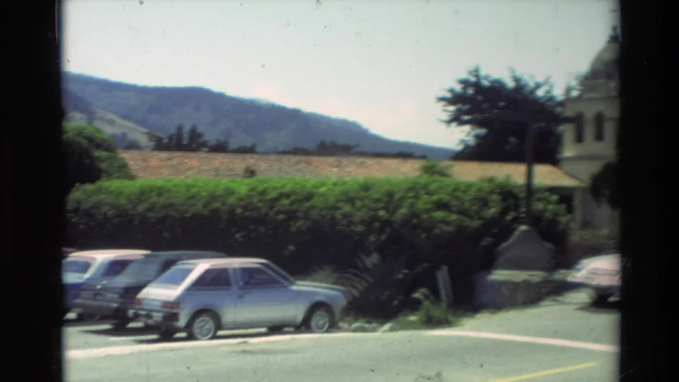1982 Carmel Mission area view showing parked cars, landscaped hedges, hills, and part of the basilica architecture.