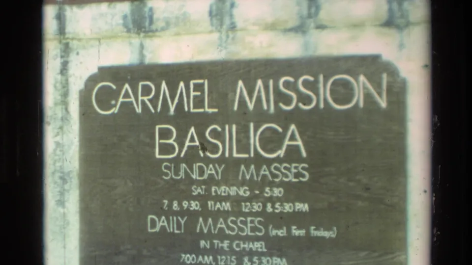 Close view of a Carmel Mission Basilica sign with Sunday and daily mass times, filmed on 8mm home-movie stock.