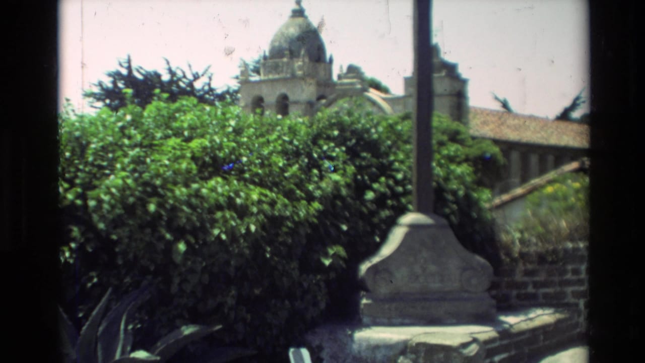 1982 8mm frame showing a posted mass-times sign with greenery and mission grounds nearby in Carmel, California.