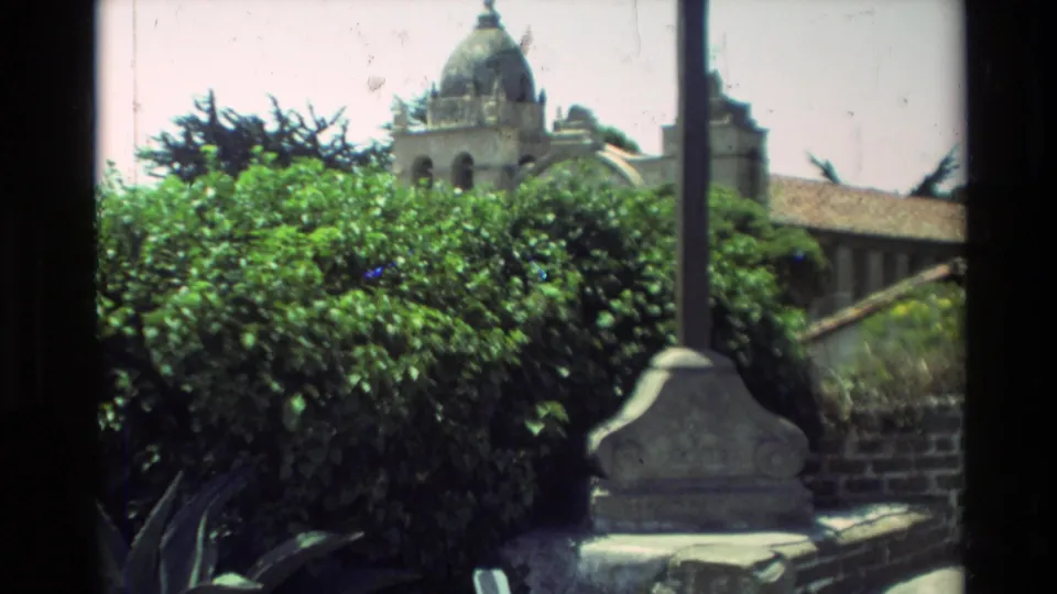 1982 8mm frame showing a posted mass-times sign with greenery and mission grounds nearby in Carmel, California.