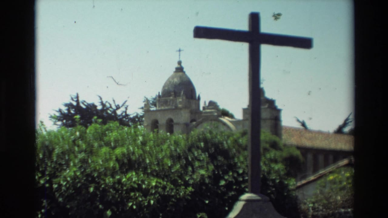 Mission building and stone cross seen above hedges.