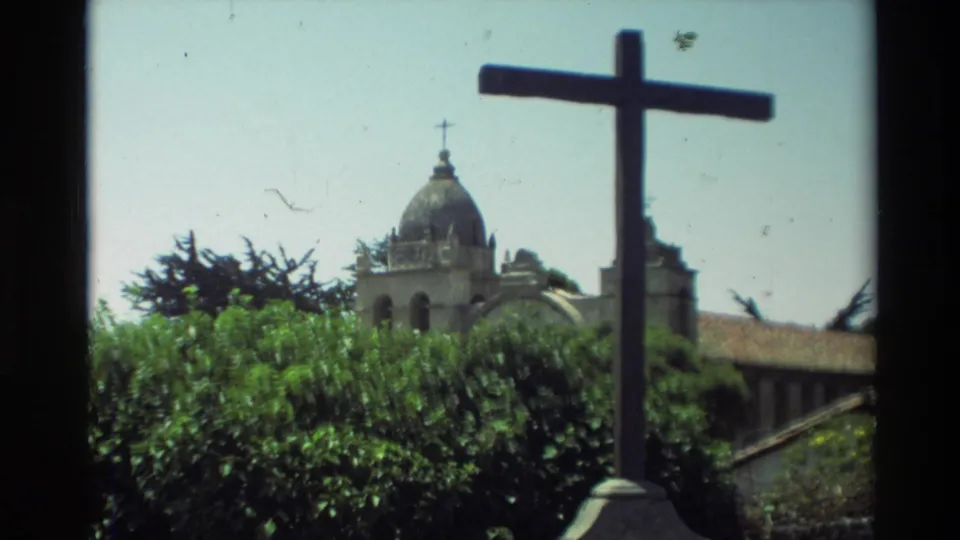 Carmel Mission Basilica exterior with a foreground stone cross, hedges, dome, and bright sky in 1982 home-movie footage.