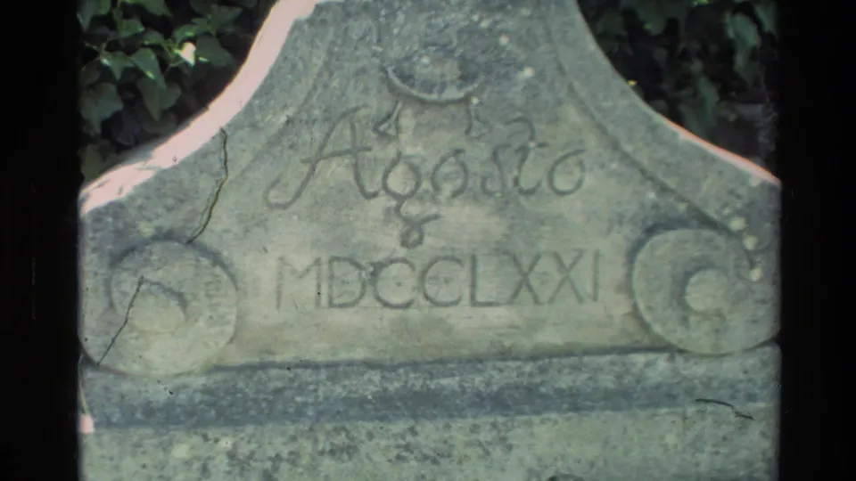 Close 1982 8mm view of a carved stone marker at Carmel Mission, showing decorative lettering and Roman numerals.