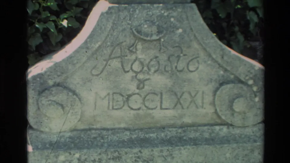 Close-up of a weathered carved stone marker at Carmel Mission, with lettering and Roman numerals visible on 1982 8mm film.