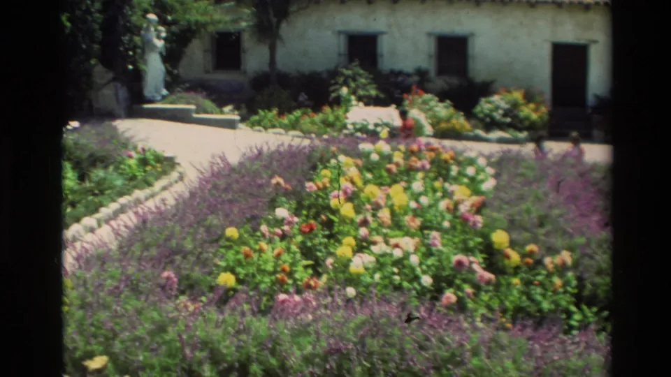 Carmel, California 1982 frame of a sunlit courtyard garden with colorful flowers, purple planting, white stucco walls, and a distant statue.