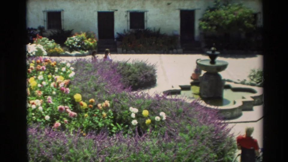 Carmel, California 1982 frame showing a landscaped courtyard with flower beds, white stucco walls, a stone fountain, and visitors at a distance.