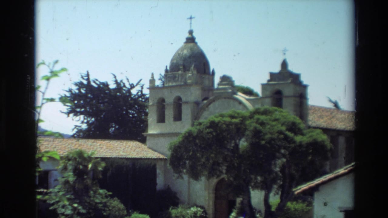 1982 Carmel, California home-movie view of a Spanish-style mission exterior with a domed bell tower, crosses, tile roof, and surrounding trees.