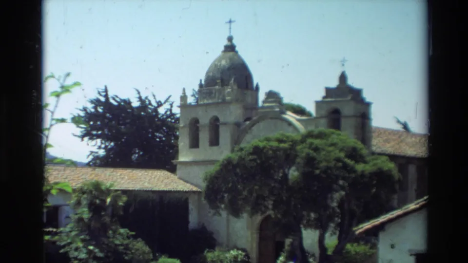 1982 Carmel, California home-movie view of a Spanish-style mission exterior with a domed bell tower, crosses, tile roof, and surrounding trees.