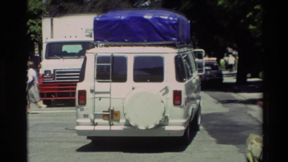 White van carrying roof cargo on a tree-lined Carmel street, with nearby vehicles and a pedestrian at the edge of the frame.