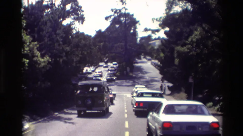 1982 Carmel street view with cars moving along a tree-lined road, including a van and several sedans in bright sun and shade.