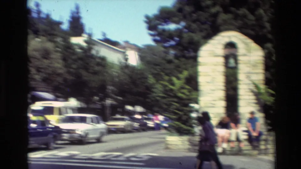 Carmel street corner with pedestrians near a distinctive stone structure, parked vehicles, and trees in a 1982 home-movie view.